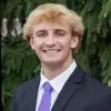 Smiling young man with blond hair wearing a dark suit and purple tie in front of green foliage.