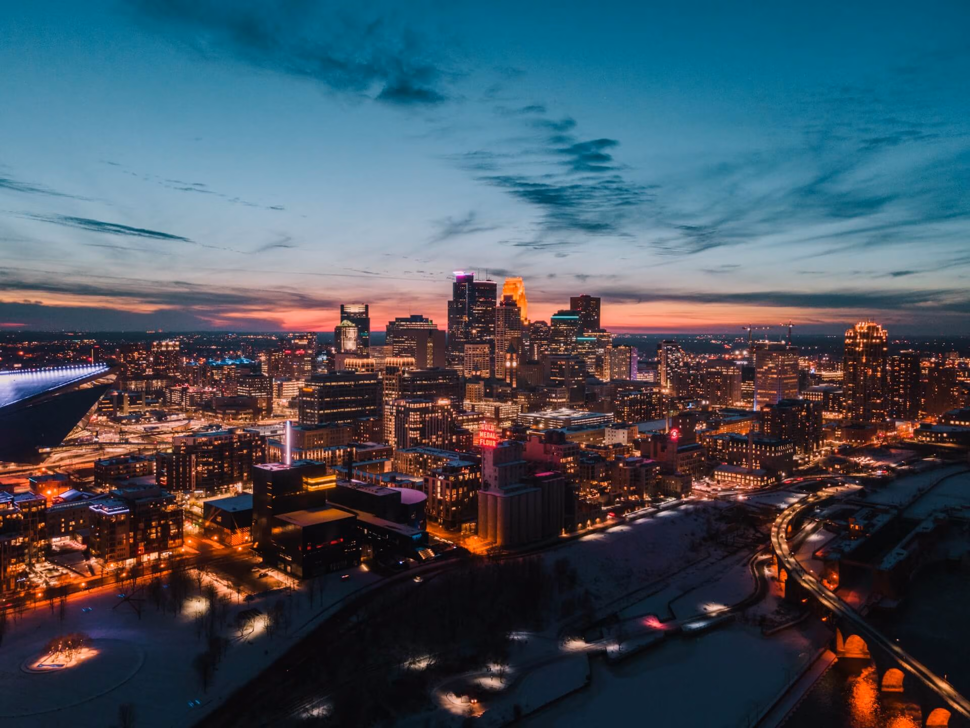Aerial view of Minneapolis city skyline at dusk with illuminated buildings and a colorful sunset sky.